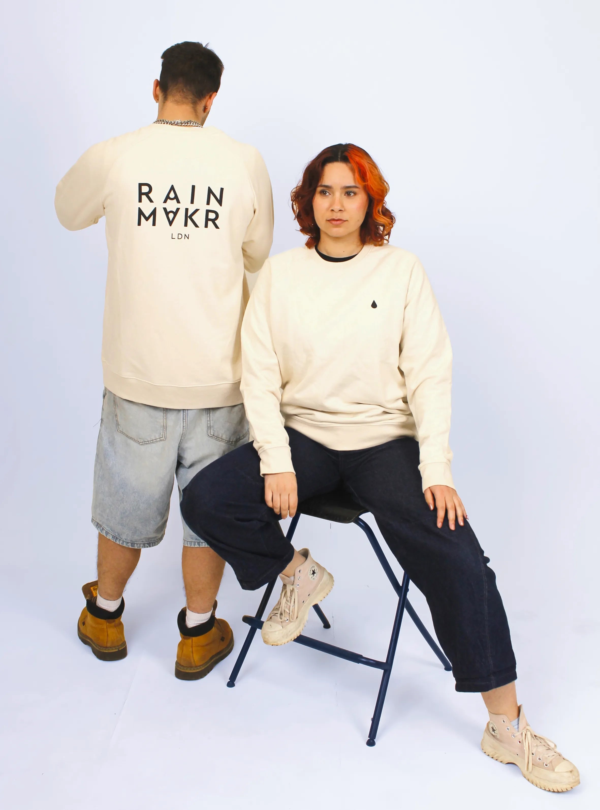 Two models wearing a beige long-sleeve crewneck jumper with 'RAINMAKR' text printed at the middle back.